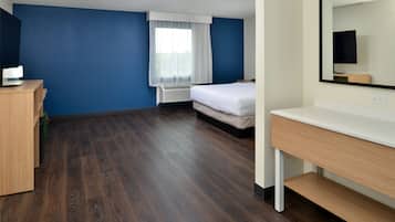 A hotel room with a wooden floor, a bed, a desk, a chair, a mirror, and a window with curtains.