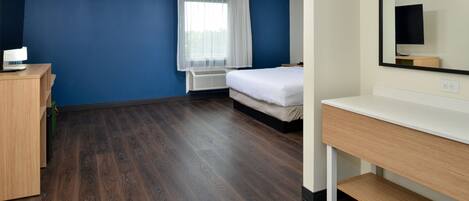A hotel room with a wooden floor, a bed, a desk, a chair, a mirror, and a window with curtains.