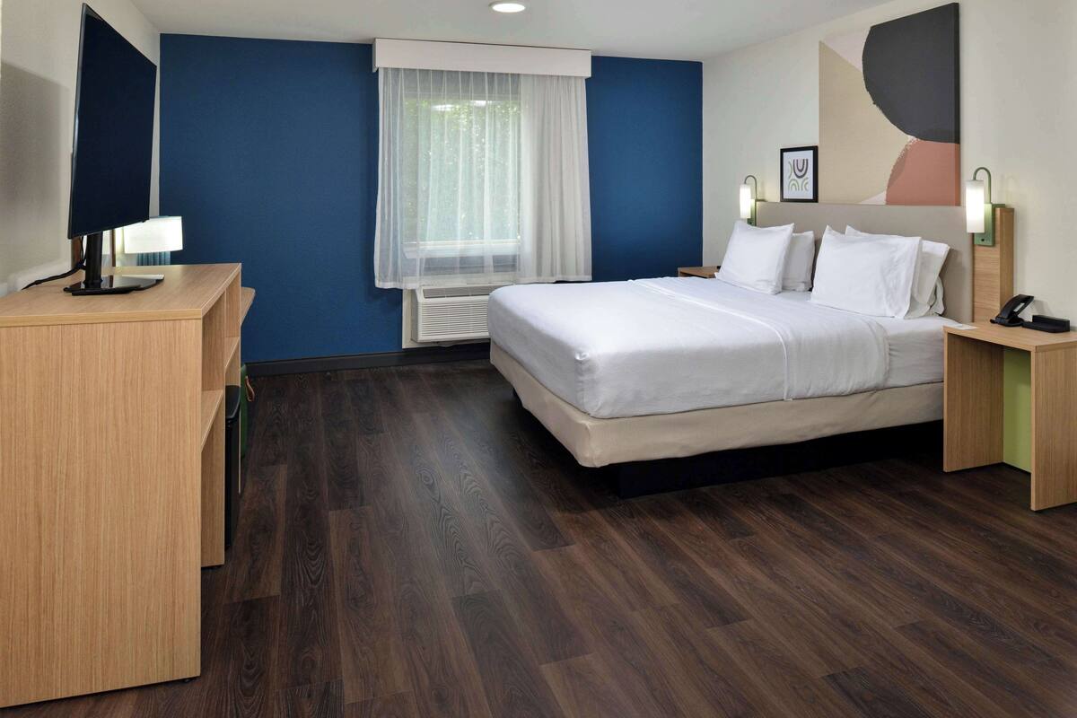 A hotel room with a bed, bedside table, lamp, and a desk with a computer monitor.