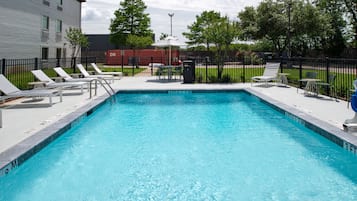 A rectangular swimming pool with clear blue water, surrounded by a black fence. There are lounge chairs and a table with an umbrella on the poolside.