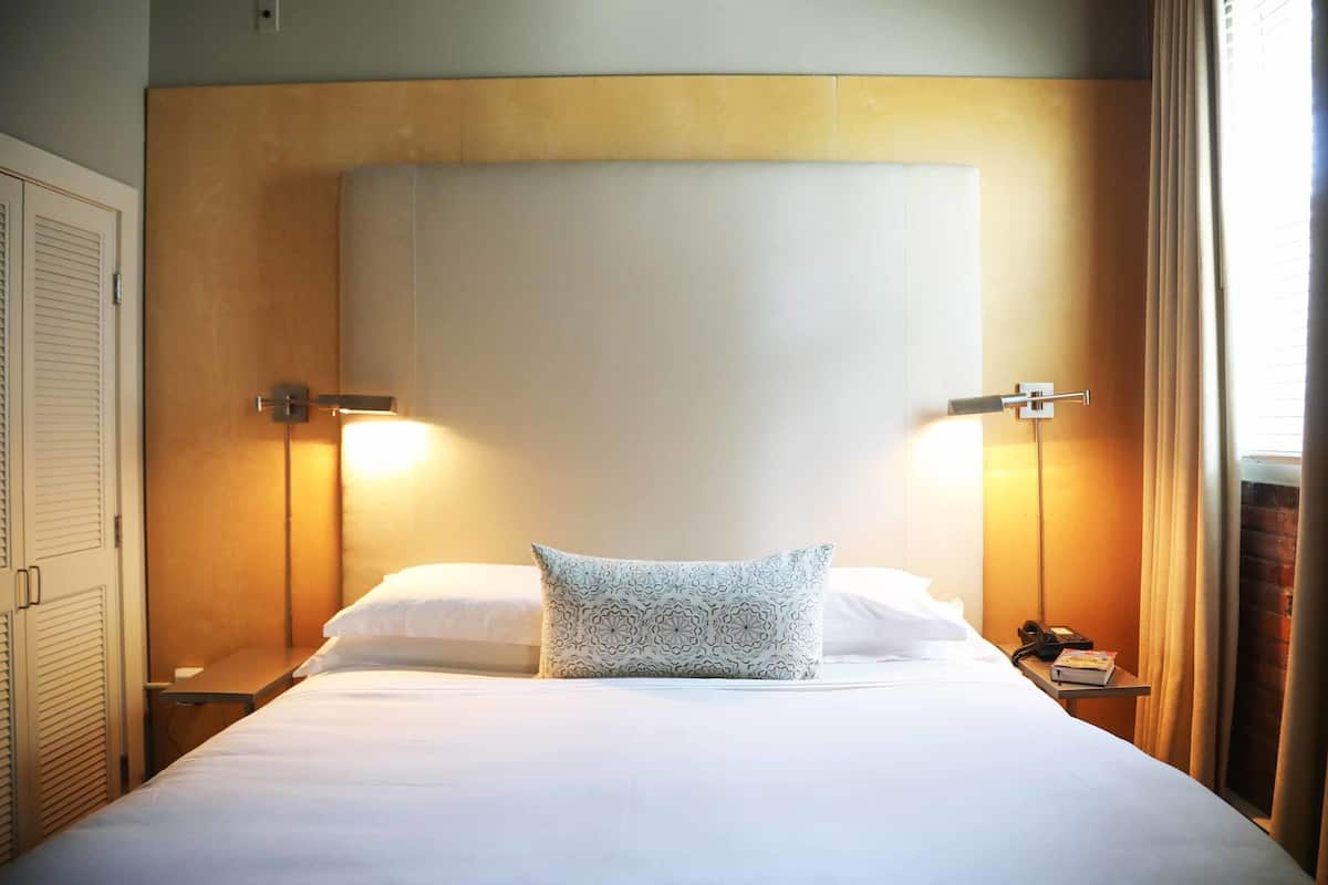 A hotel room with a large bed, a bedside table with a lamp, a telephone, and a window with curtains.