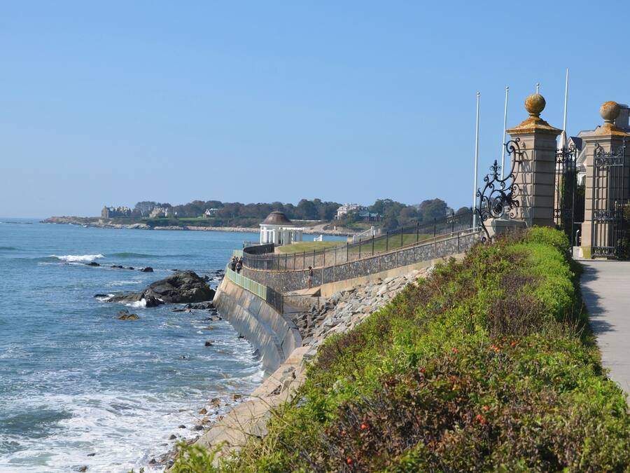 Un paseo marítimo con un muro de piedra, un sendero y vistas al mar y edificios a lo lejos.