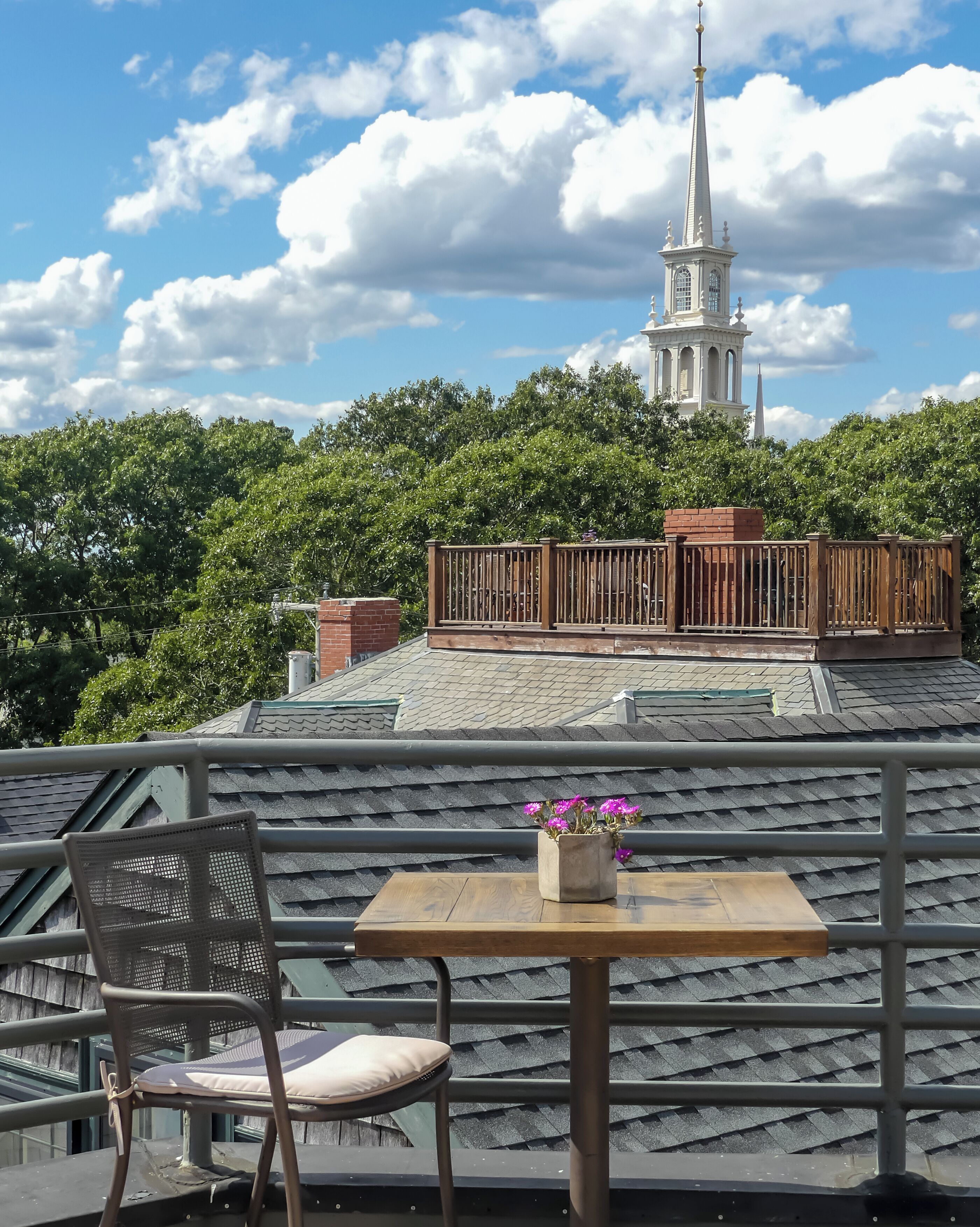A balcony with a table and chair, a flower pot, and a view of a church spire.