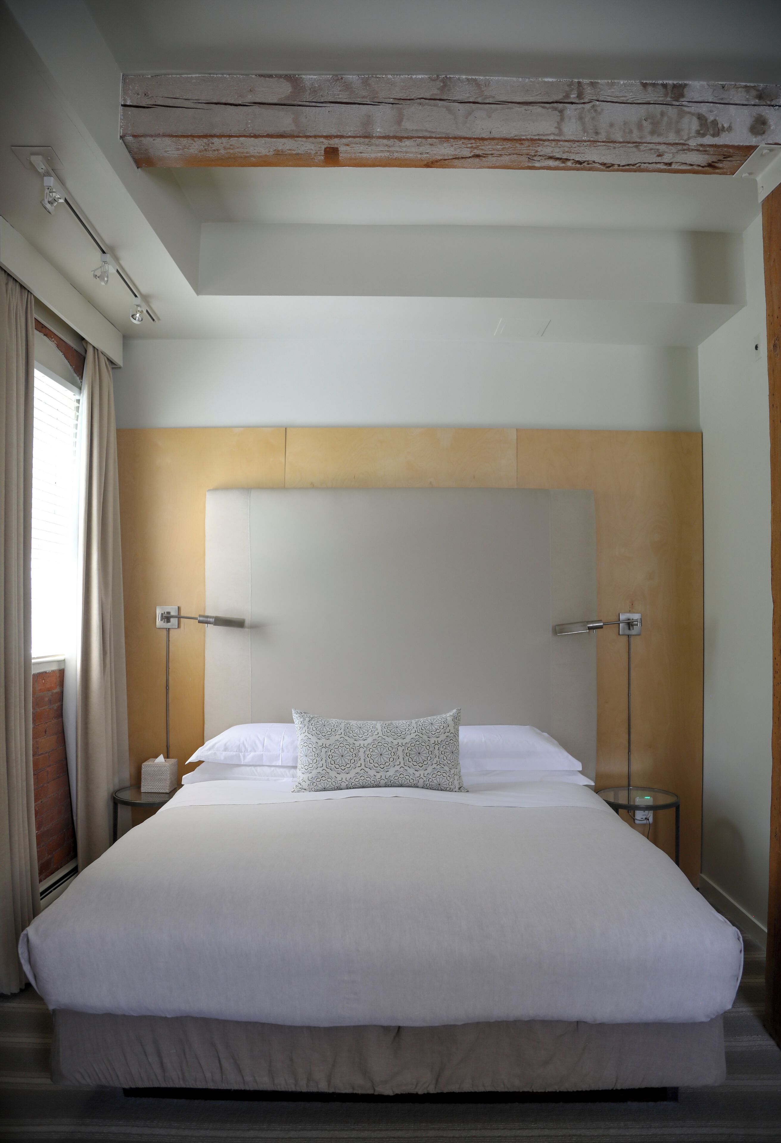 Uma cama grande com lençóis brancos, cabeceira de madeira, abajures e uma janela com cortinas.