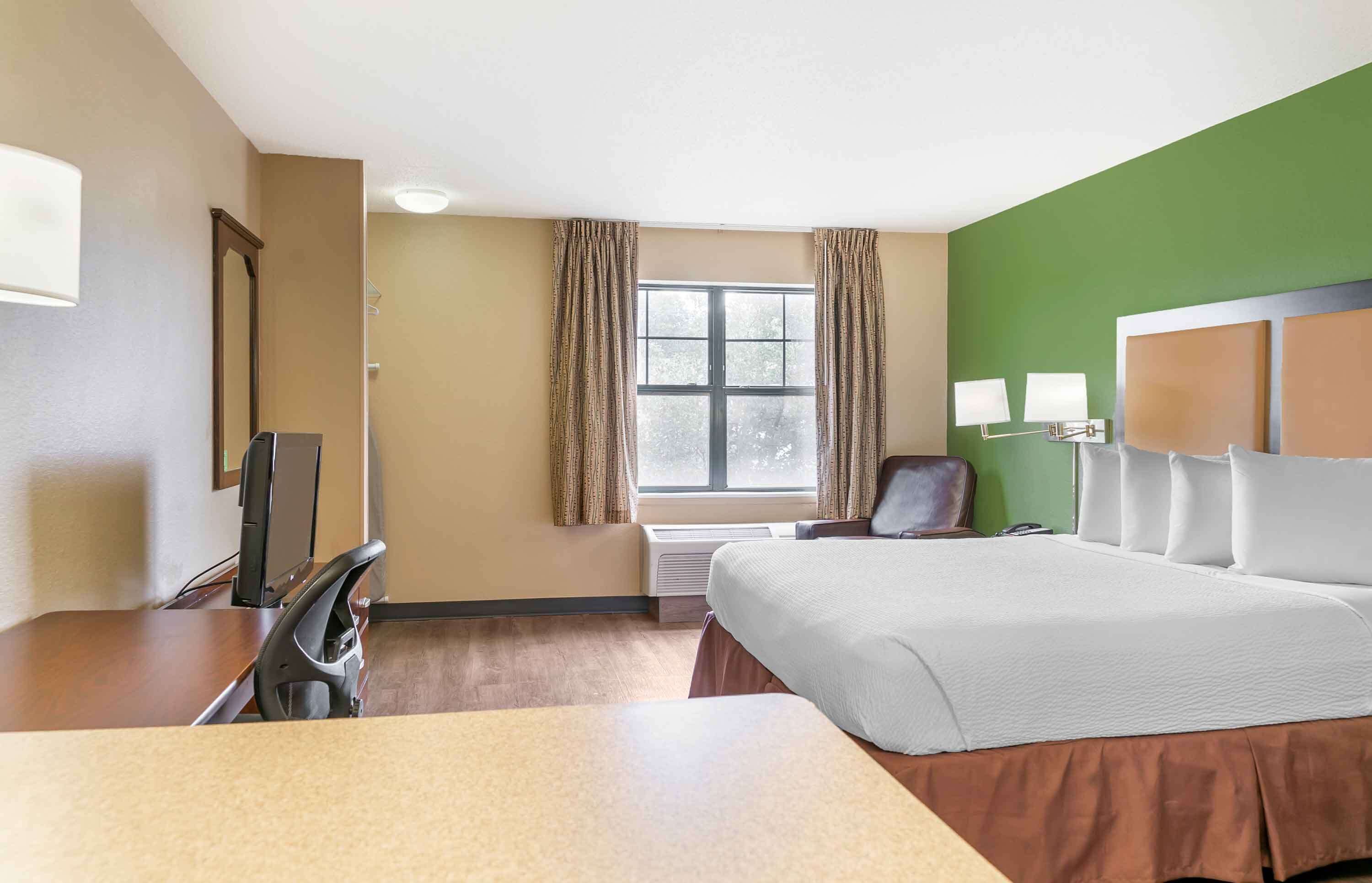 A hotel room with a bed, a desk, a chair, a TV, a window with curtains, and a green accent wall.