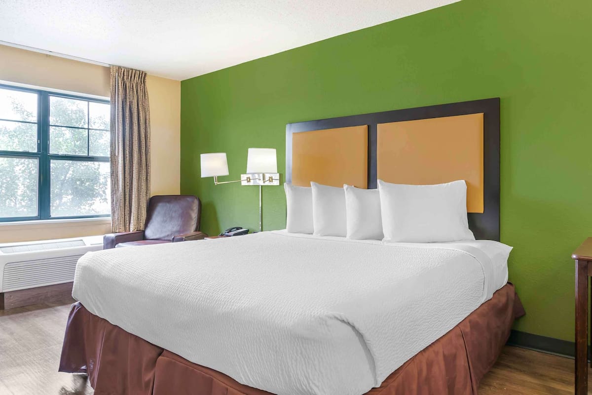 Camera d'albergo con un letto grande, pareti verdi, una finestra con tende, una sedia e un comodino con lampade.