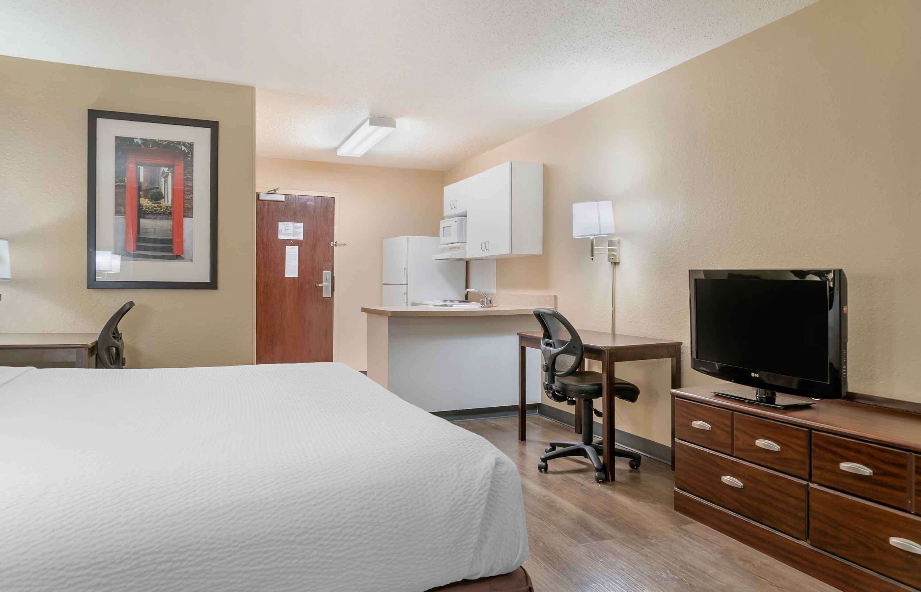 Una habitación de hotel con cama, escritorio, silla, televisión y kitchenette.