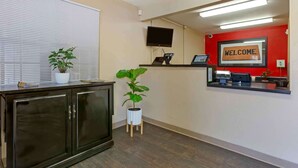 Ein Empfangsbereich mit einem dunklen Holzschrank, zwei Topfpflanzen, einer roten Wand mit einem 'WELCOME'-Schild, einem Wand-Fernseher und einem Schreibtisch mit Computer und Telefon.