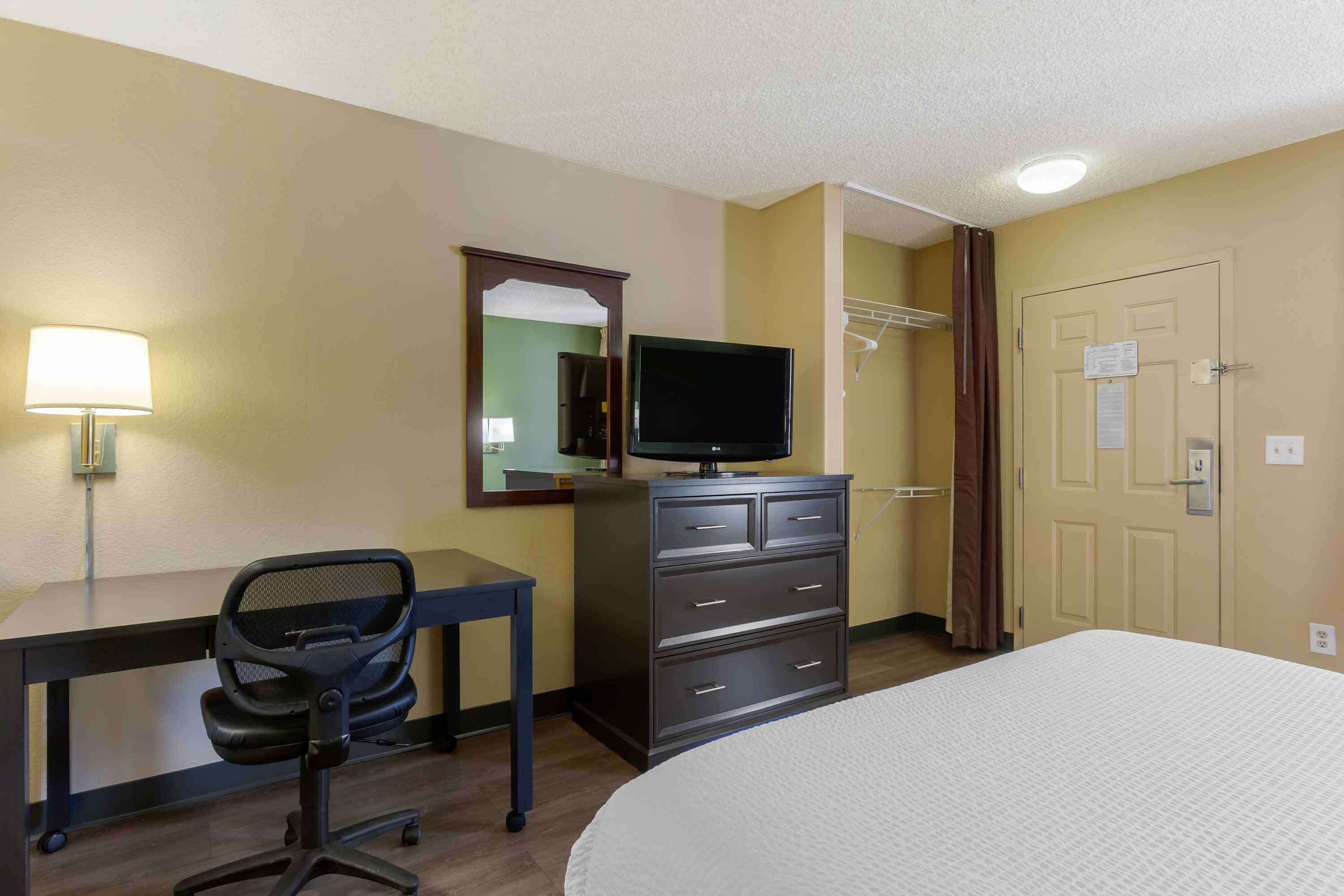 A hotel room with a bed, a desk with a chair, a television on a dresser, and a door with a notice.
