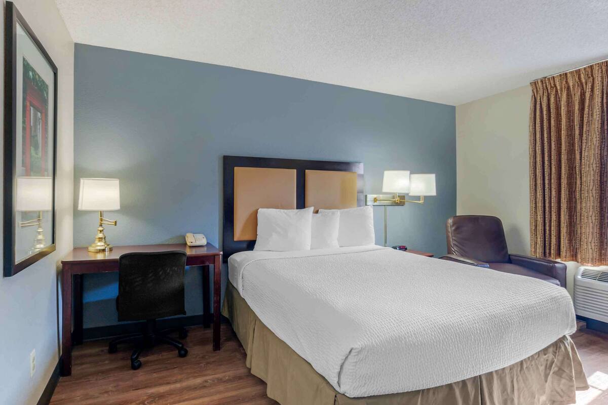 Une chambre d’hôtel avec un grand lit, un bureau avec une lampe, une chaise et une fenêtre avec des rideaux.