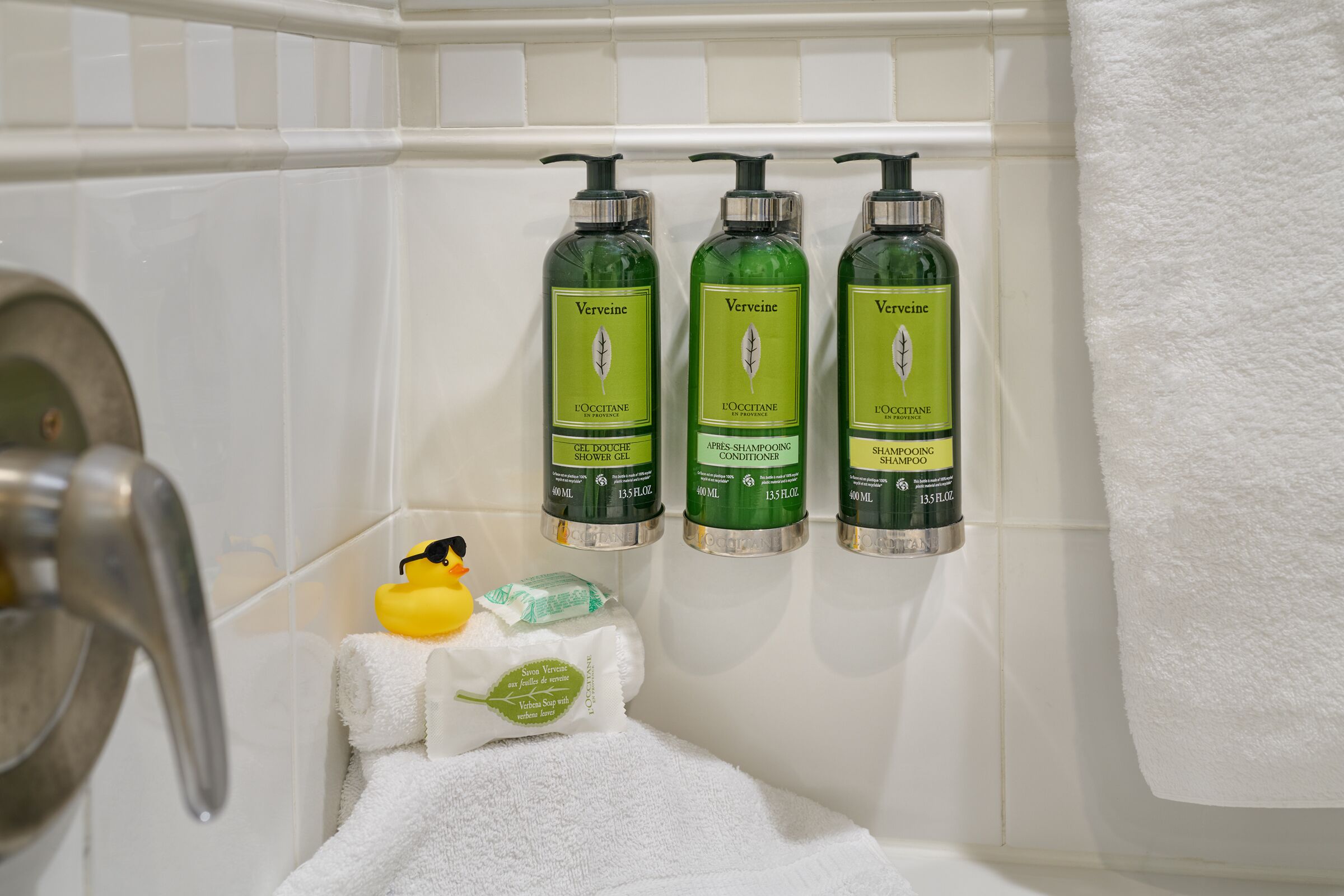 Un baño con tres botellas de gel de ducha, acondicionador y champú Verveine, una toalla blanca y un patito de goma.