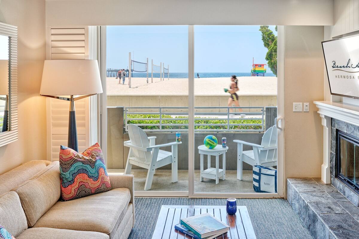 Un salon comprenant un canapé, une lampe, une table basse avec des livres, une cheminée et une vue sur la plage, où se trouve également un filet de volley-ball.