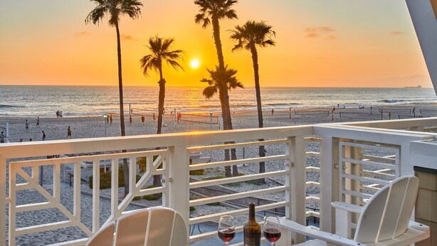Una terraza de madera con dos sillas blancas y una mesita, con vista a una playa con palmeras y un atardecer.