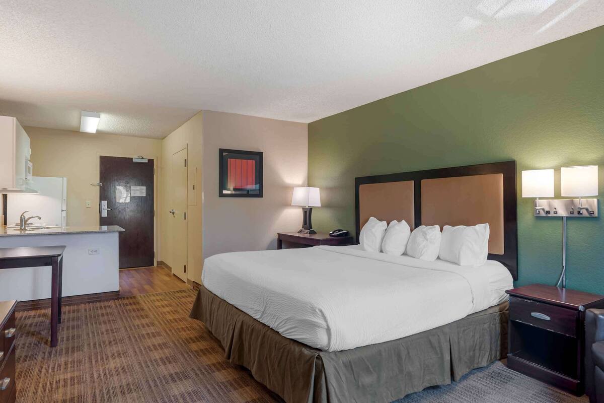 Une chambre d’hôtel avec un grand lit, un bureau avec une chaise, une petite table, une table de chevet avec une lampe et un téléviseur fixé au mur.