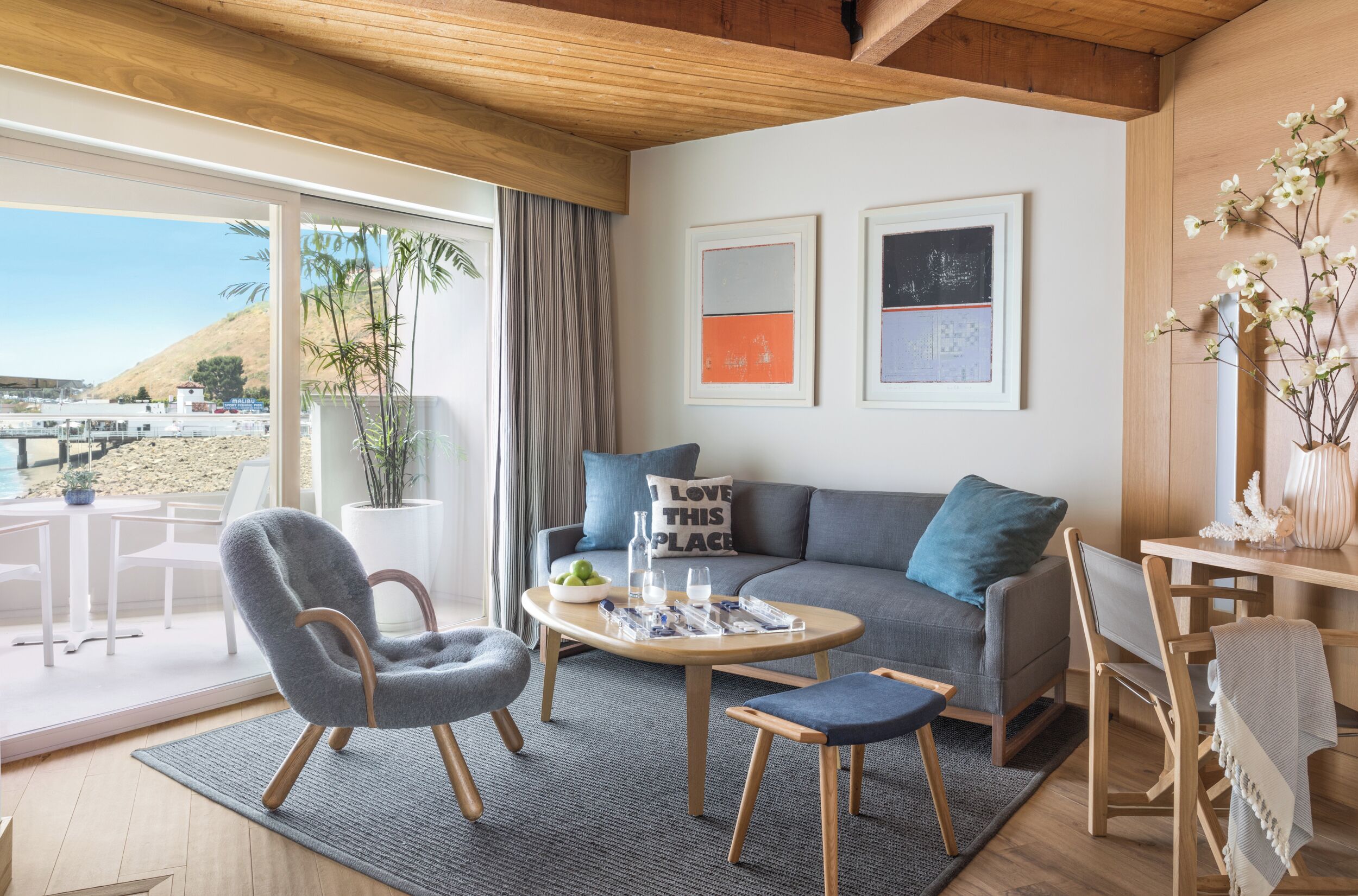 A modern living room with a grey sofa, wooden coffee table, and a view of the beach through large windows.