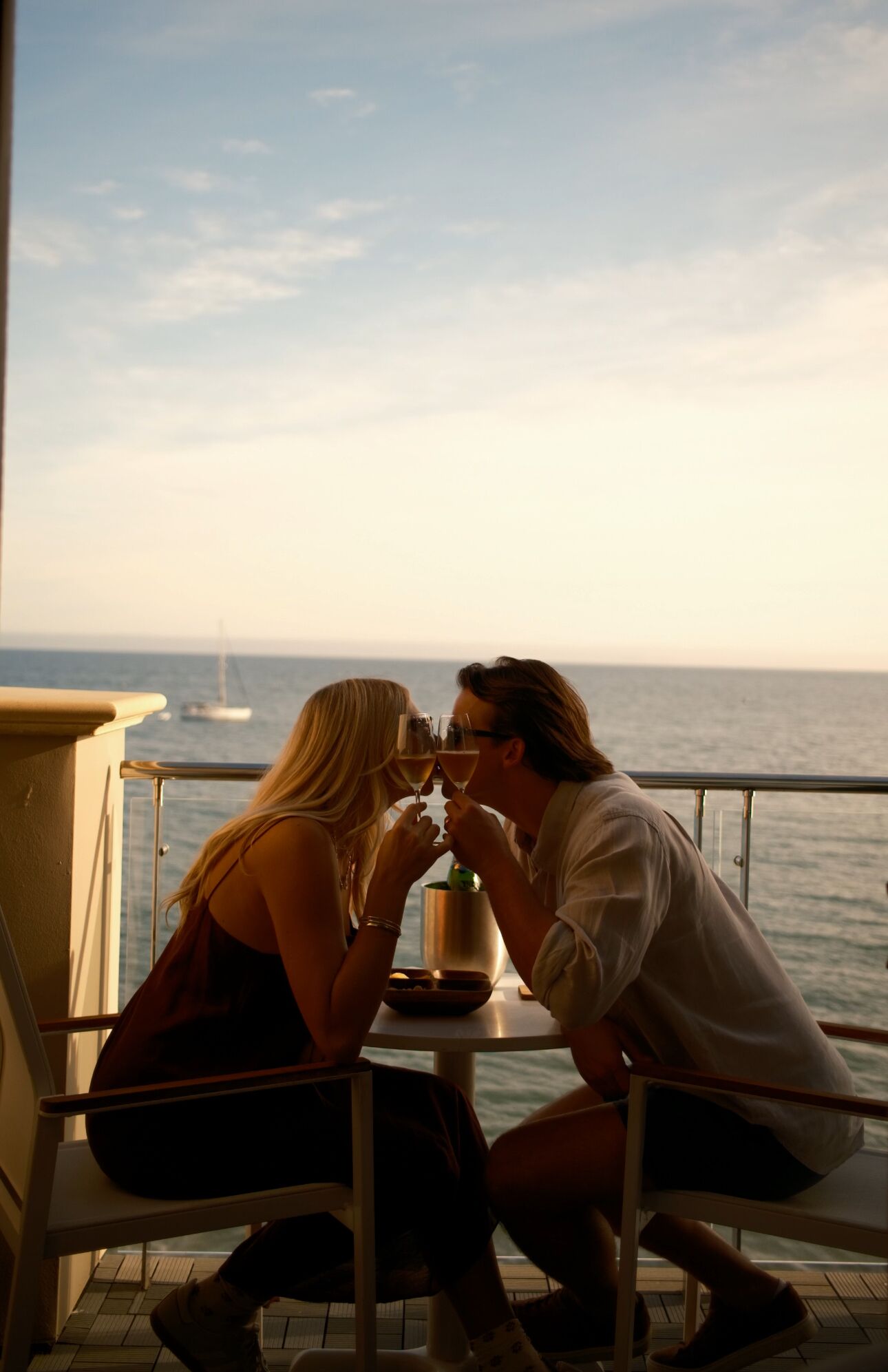 A couple sitting at a table with a view of the sea and a sailboat in the distance.