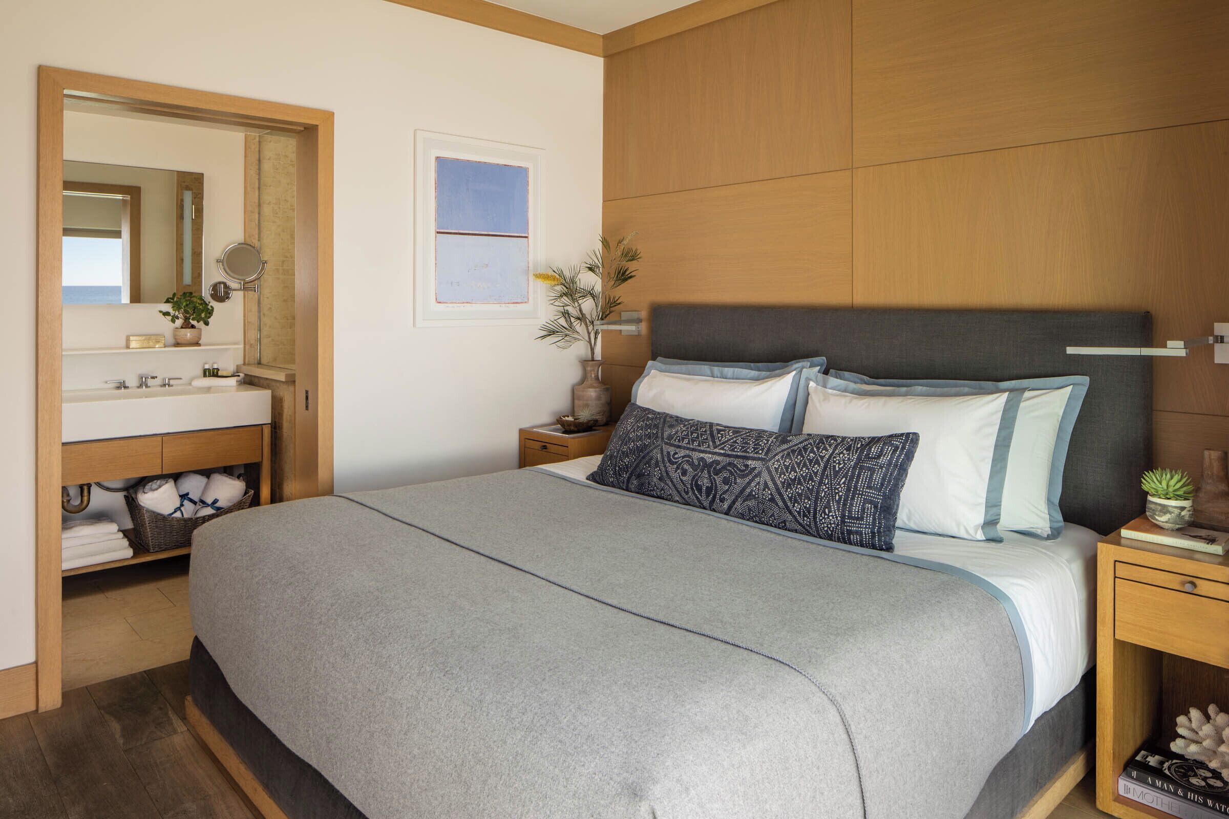 A bedroom with a large bed, a wooden headboard, a nightstand with a lamp, and a bathroom with a mirror and sink.