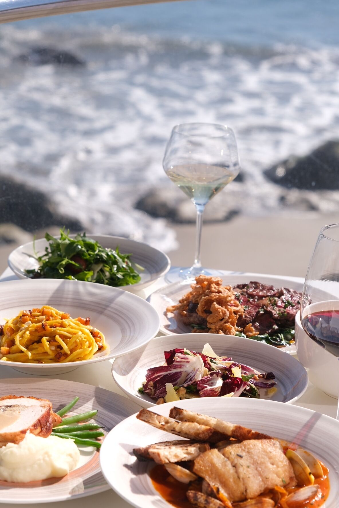 A table with various dishes and a glass of white wine, set against a backdrop of ocean waves.