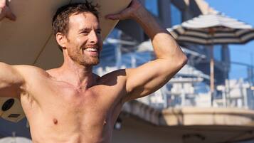 Um homem segurando uma prancha de surf com um logotipo, em área externa com guarda-sóis e um edifício ao fundo.