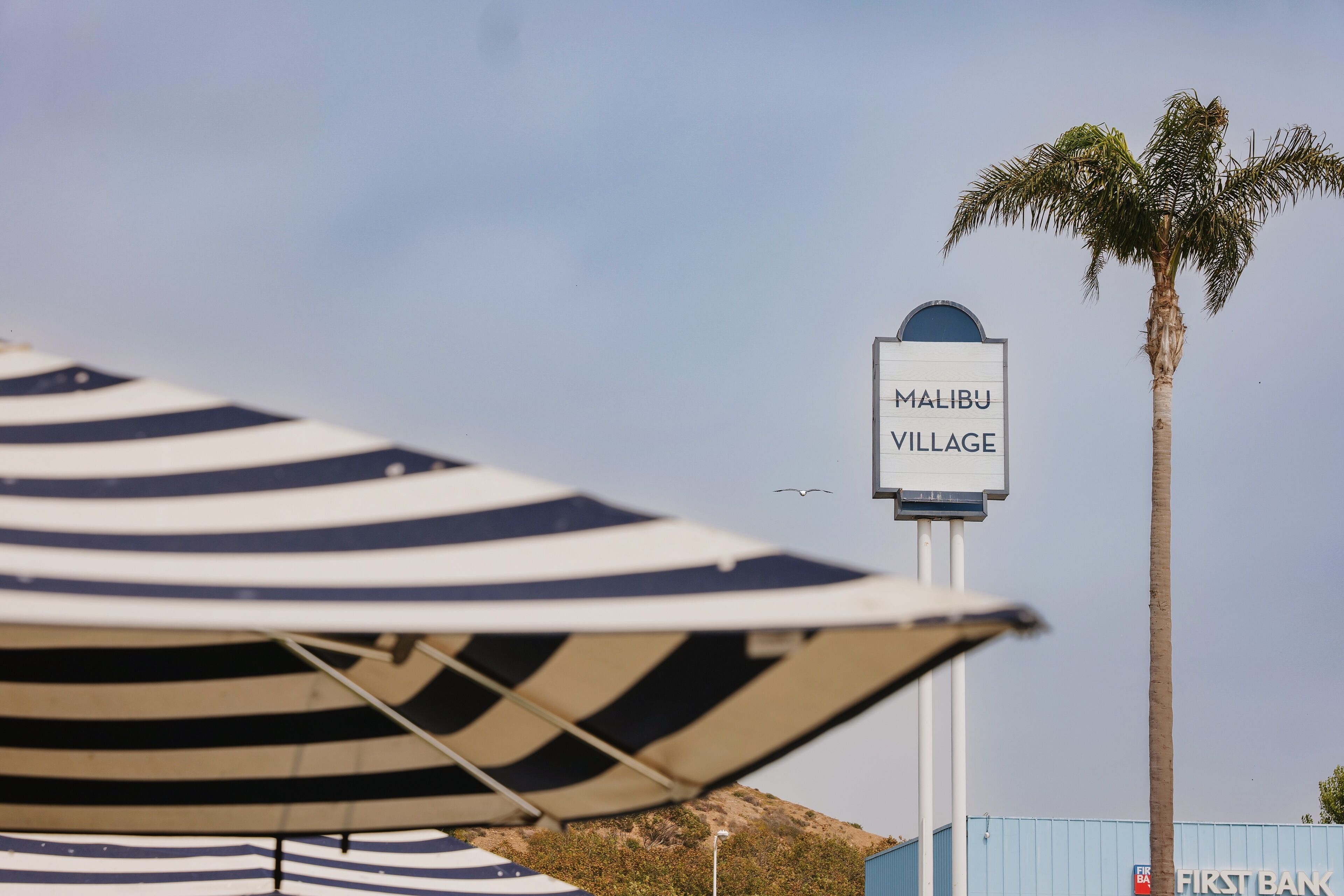 A sign for Malibu Village with a palm tree and a striped umbrella in the foreground.