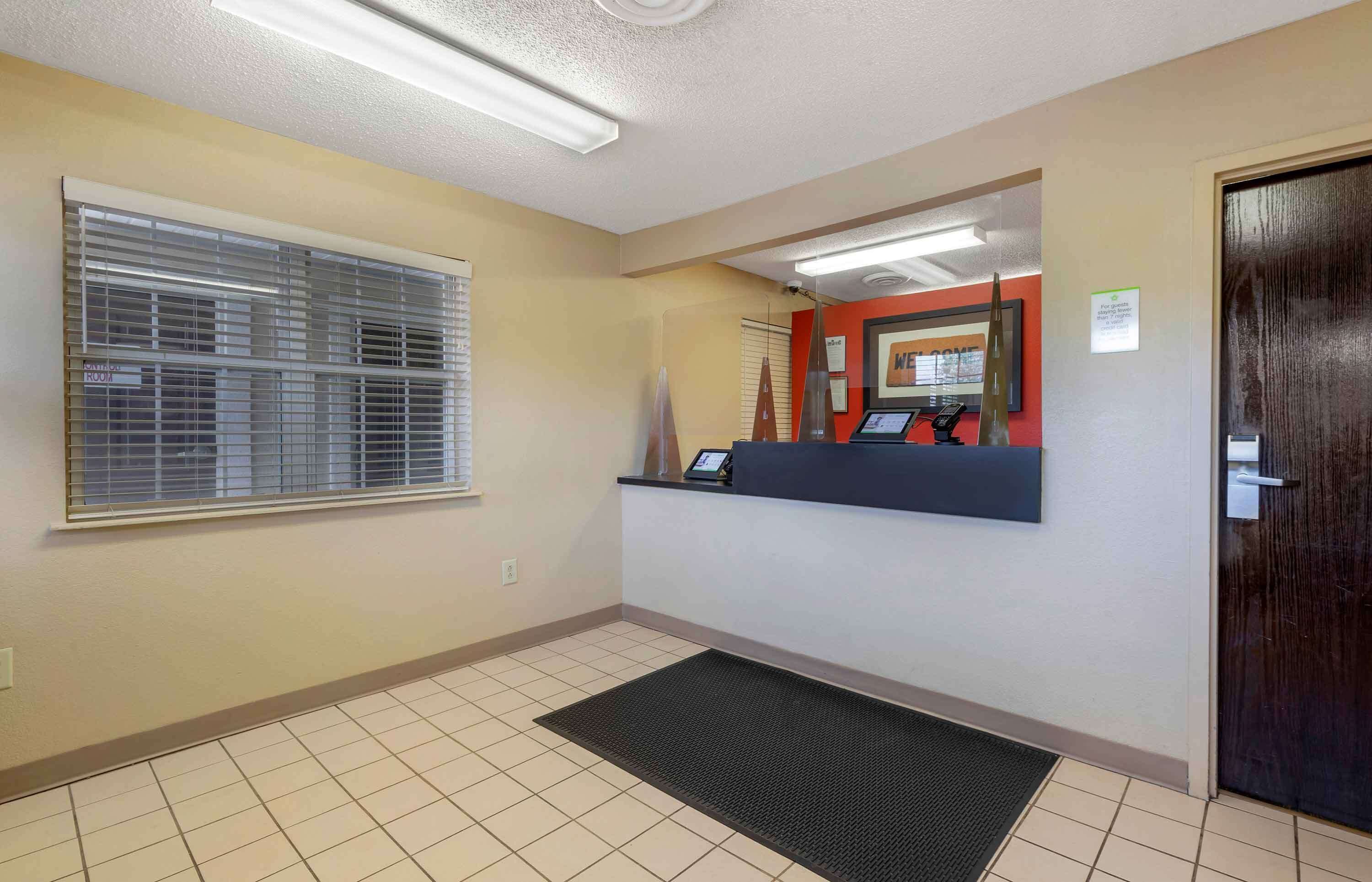 A reception area with a reception desk, a black mat at the entrance, a window with blinds, and a door.