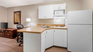 A compact kitchen with white cabinets, a microwave, a refrigerator, a sink, a wooden desk, a chair, and a television. - Extended Stay America Suites Fort Wayne South (Fort Wayne)