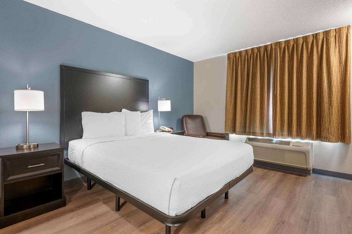 A hotel room with a bed, nightstands, a chair, a window with curtains, and an air conditioning unit.