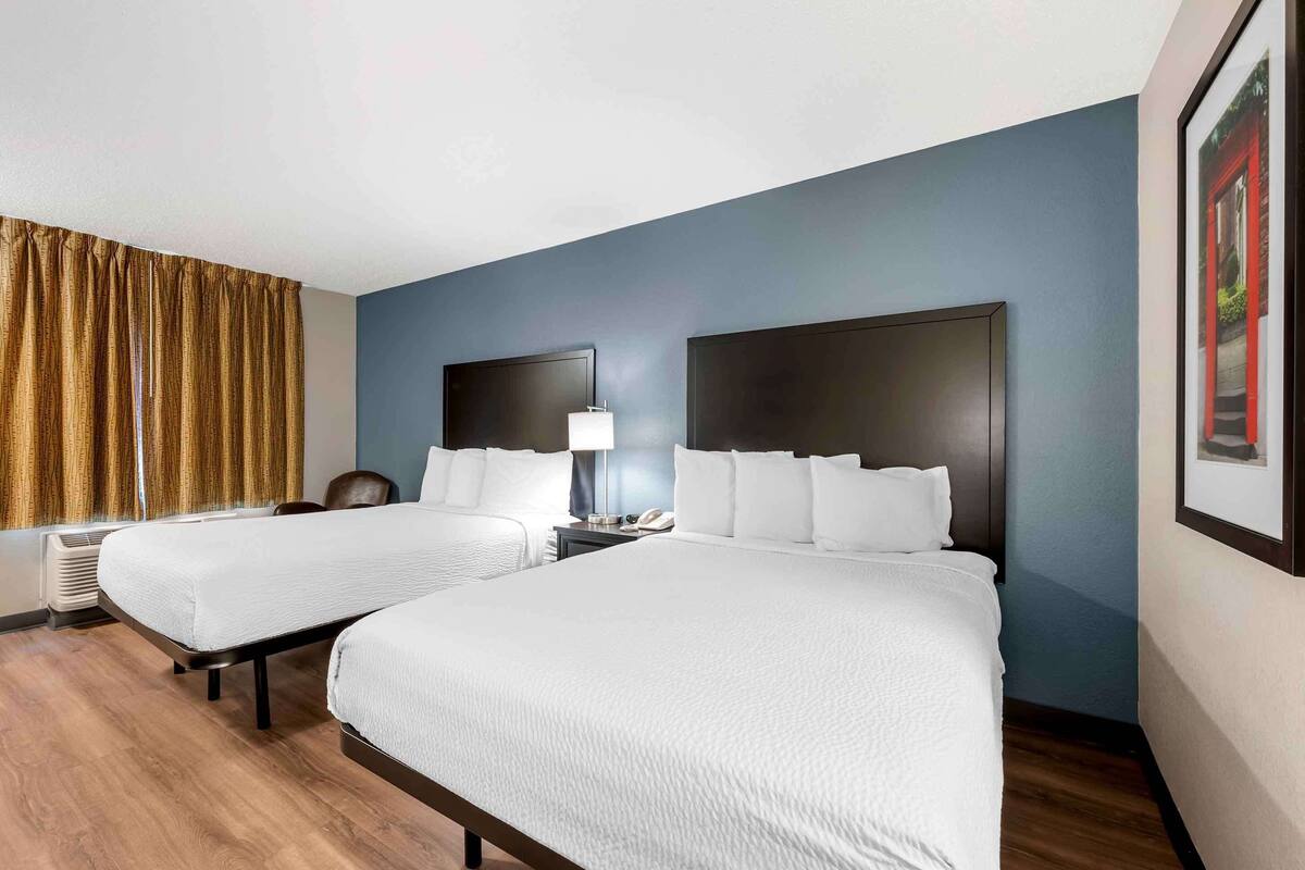 A hotel room with two beds, a nightstand with a lamp, a window with curtains, and a framed picture on the wall.