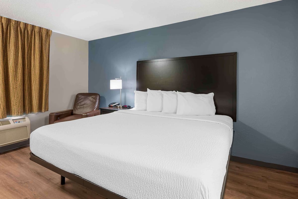 A hotel room with a large bed, a headboard, a chair, a desk with a lamp, and an air conditioning unit.