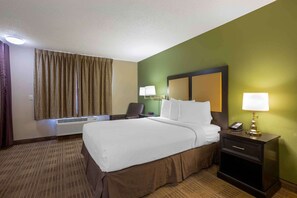 A hotel room with a large bed, a bedside table with a lamp, a chair, and a desk with a telephone.