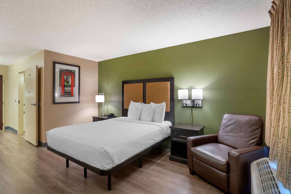 Habitación de hotel con una cama, mesitas de noche, una silla y una lámpara fijada a la pared.