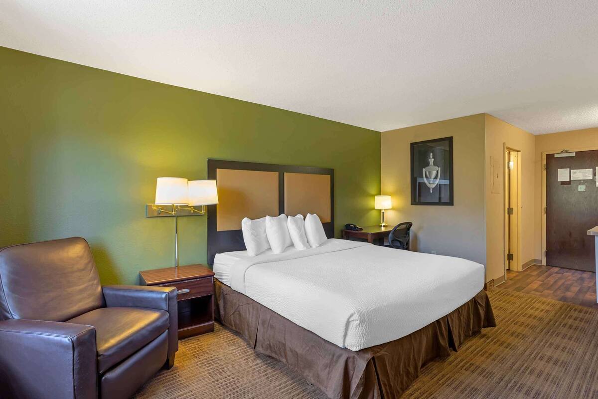Habitación de hotel con una cama grande, una silla, una mesita de noche, una lámpara y una puerta.