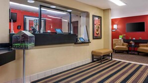 Zona de recepción de un hotel con un mostrador, un espejo grande, un área de estar con dos sillas y un banco, y suelo alfombrado.