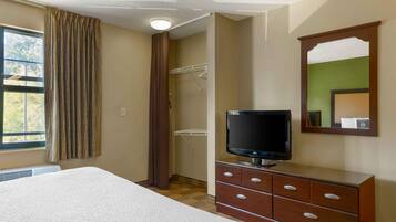 A hotel room with a bed, a TV on a wooden dresser, a closet with a hanging rack, and a window with curtains.