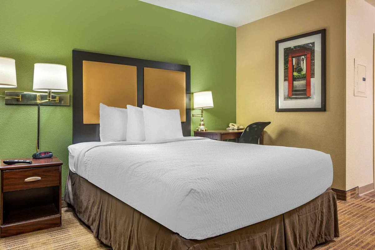 Habitación de hotel con una cama grande, dos lámparas de noche, una mesita de noche con un reloj, un escritorio con una silla y un cuadro en la pared.
