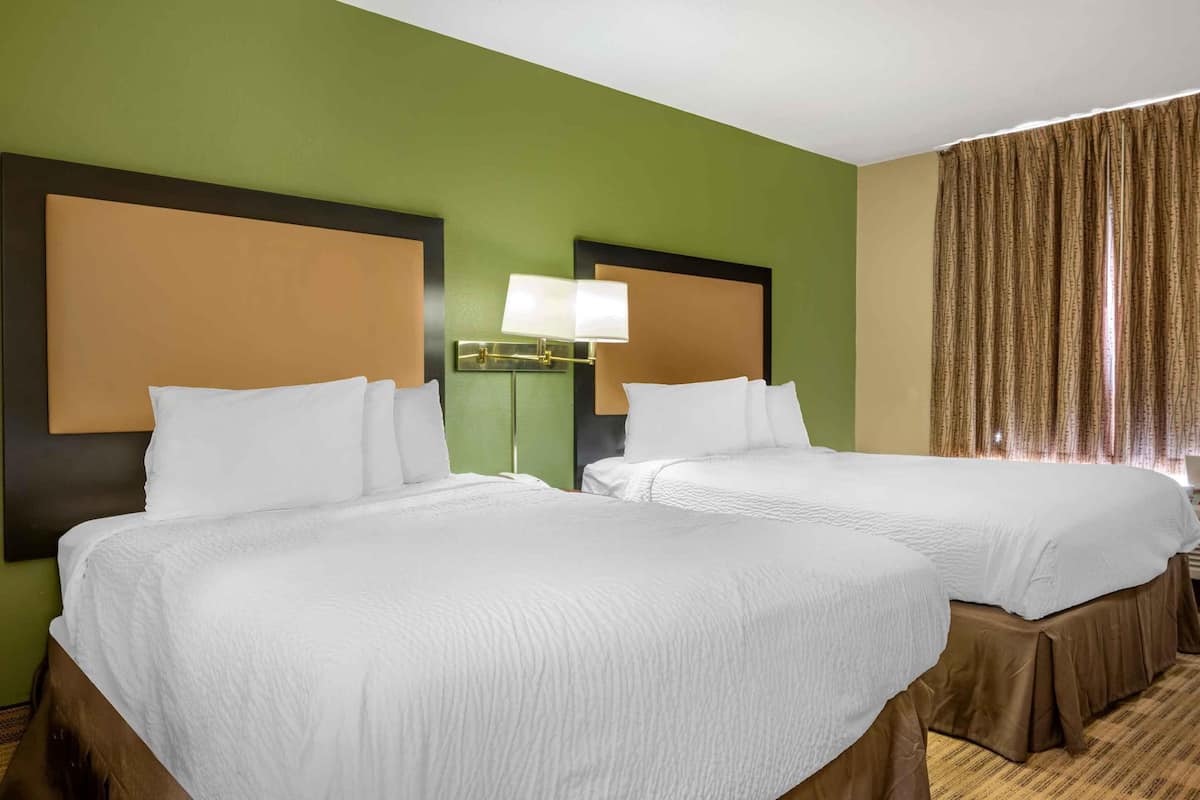 Habitación de hotel con dos camas, paredes verdes, un ventanal grande con cortinas y una lámpara de noche.