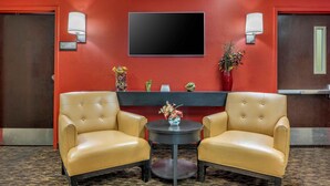 Vestíbulo de hotel con dos sillones de cuero, una mesa redonda con un jarrón de flores, un televisor de pantalla plana montado en una pared roja y un mostrador de recepción con un letrero que dice 'MECHANICAL ROOM'.