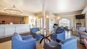 Een hotel lobby met een receptiebalie, een zithoek met blauwe bekleden stoelen, een glazen salontafel, een open haard en een groot raam.