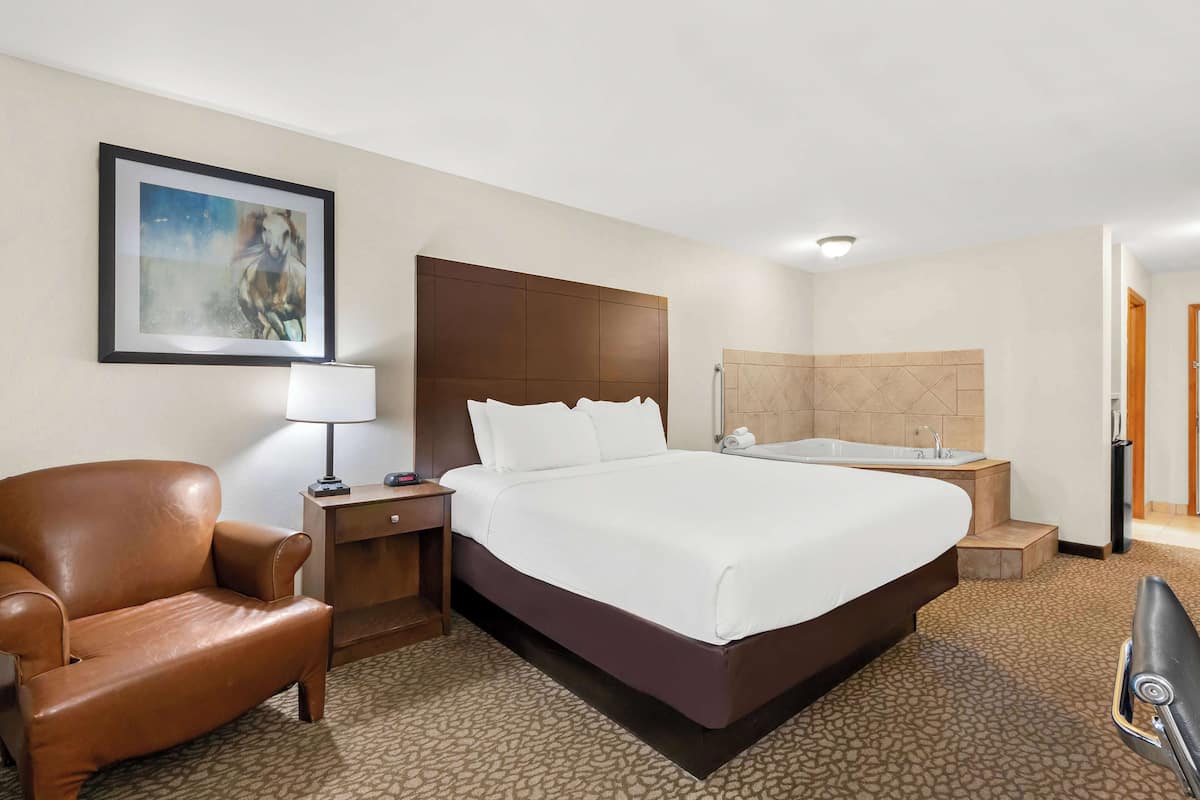 A hotel room with a large bed, a brown leather chair, a desk with a lamp, a painting on the wall, and a bathtub.