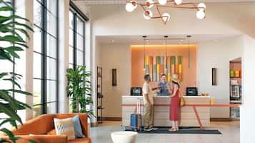 A hotel reception area with a desk, a man and a woman standing, a large window, and a plant.