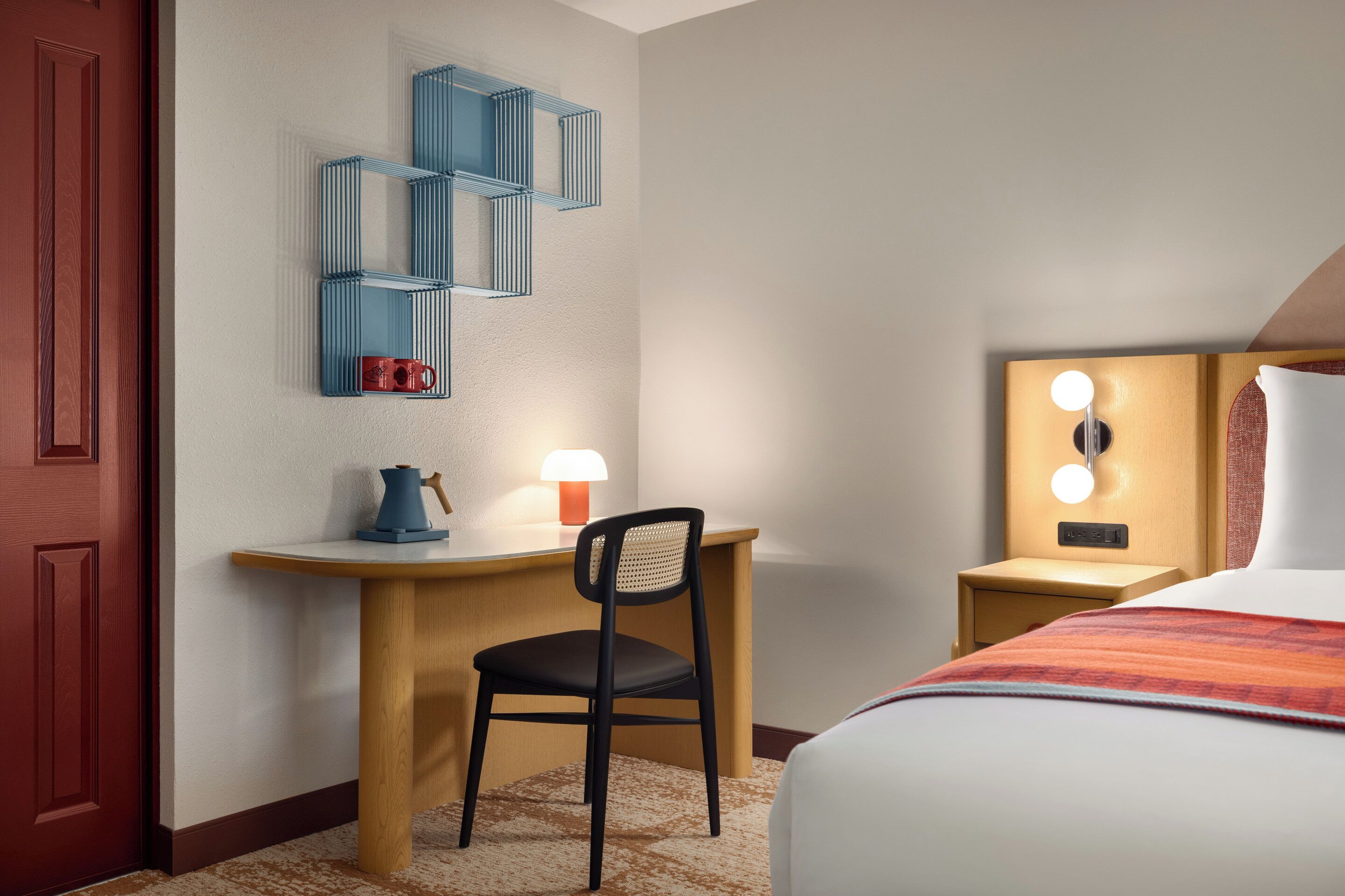 Una camera d'albergo con un letto, una scrivania con una sedia, uno scaffale a muro, un comodino con una lampada e un punto luce a parete.