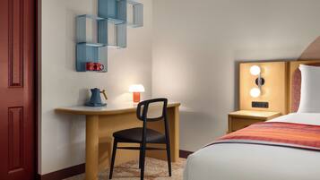Habitación de hotel con cama, escritorio y silla, estante en la pared, mesita de noche con lámpara y aplique de pared.