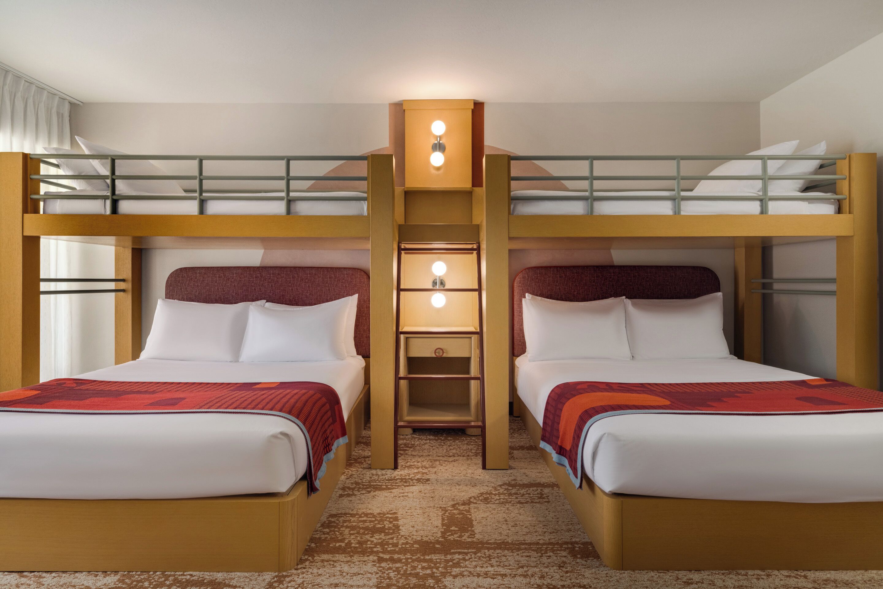 Quarto de hotel com duas camas, incluindo uma beliche, uma pequena mesa de cabeceira com abajur e piso coberto por carpete.