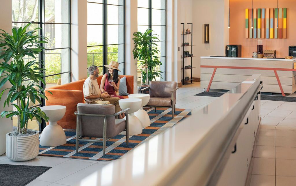 Un moderno hall de hotel con amplios ventanales, un mostrador de recepción, zona de estar con sofás y sillones, y plantas en macetas.