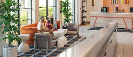 Um lobby de hotel moderno com grandes janelas, um balcão de recepção, área de estar com sofás e cadeiras, e plantas em vasos.