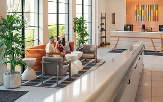 Un hall d’hôtel moderne doté de grandes fenêtres, d’un comptoir d’accueil, d’un espace salon avec des canapés et des fauteuils, et de plantes en pot.