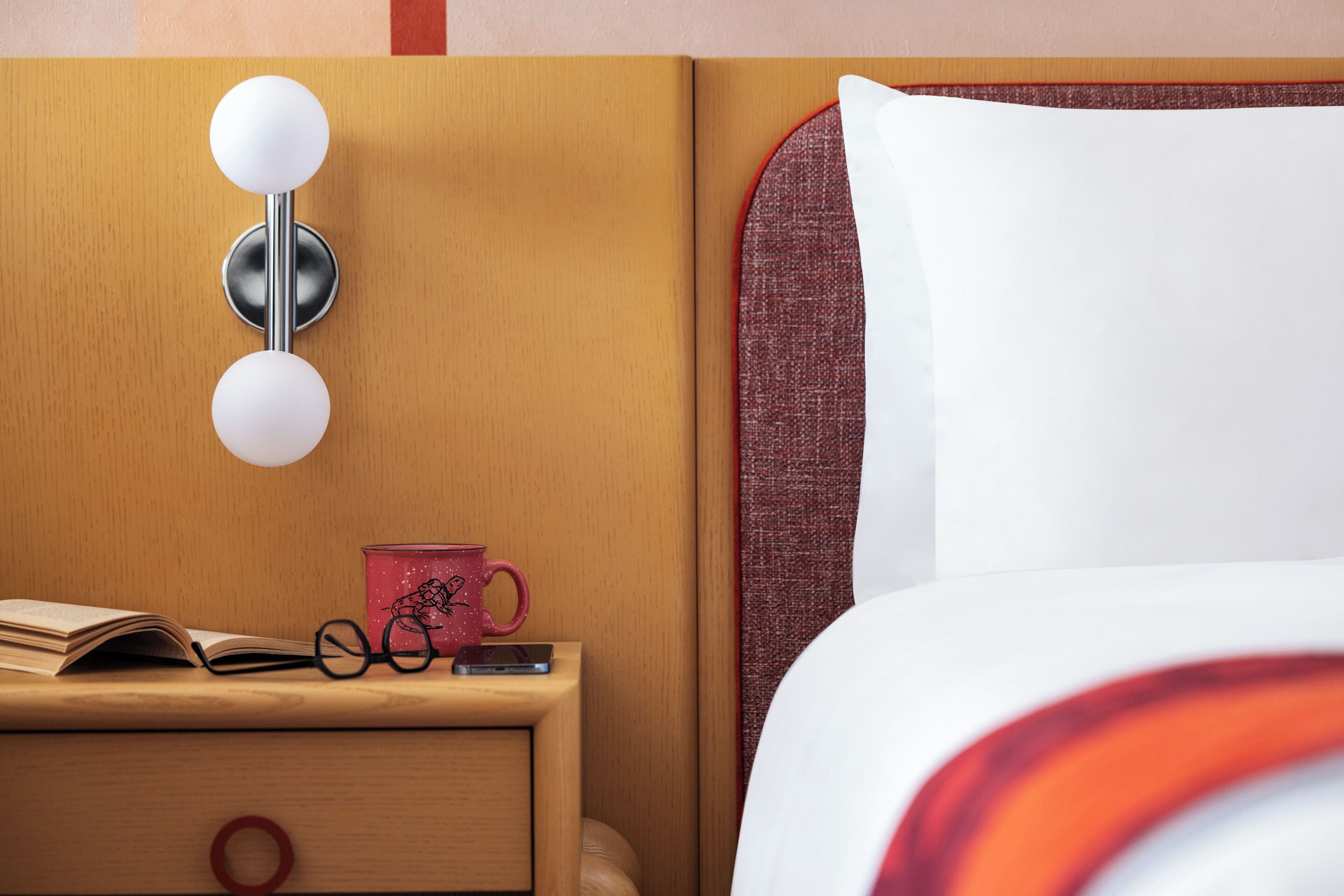 Una cama bien hecha con un almohán blanco y una manta a rayas rojas y blancas, una mesita de noche con una taza roja, un par de gafas y un smartphone, y una luminaria fijada a la pared.