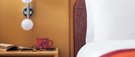 A neatly made bed with a white pillow and a red and white striped blanket, a bedside table with a red mug, a pair of glasses, and a smartphone, and a wall-mounted light fixture.