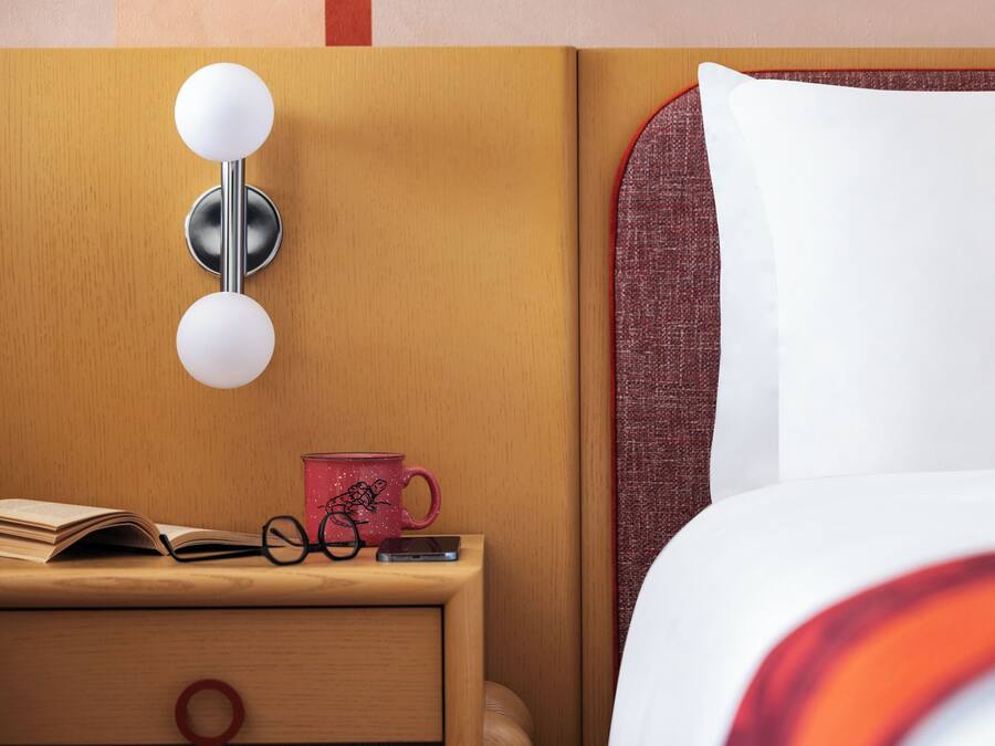 Una cama bien hecha con un almohán blanco y una manta a rayas rojas y blancas, una mesita de noche con una taza roja, un par de gafas y un smartphone, y una luminaria fijada a la pared.