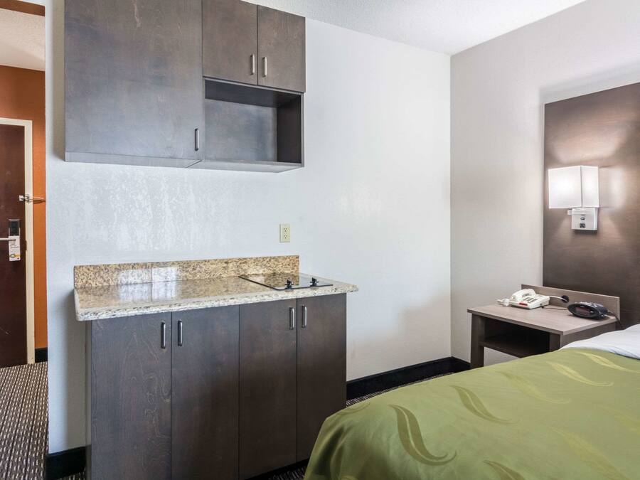 A hotel room with a bed, a small desk with a phone, a wall-mounted light, and a kitchenette with a sink and cabinets.